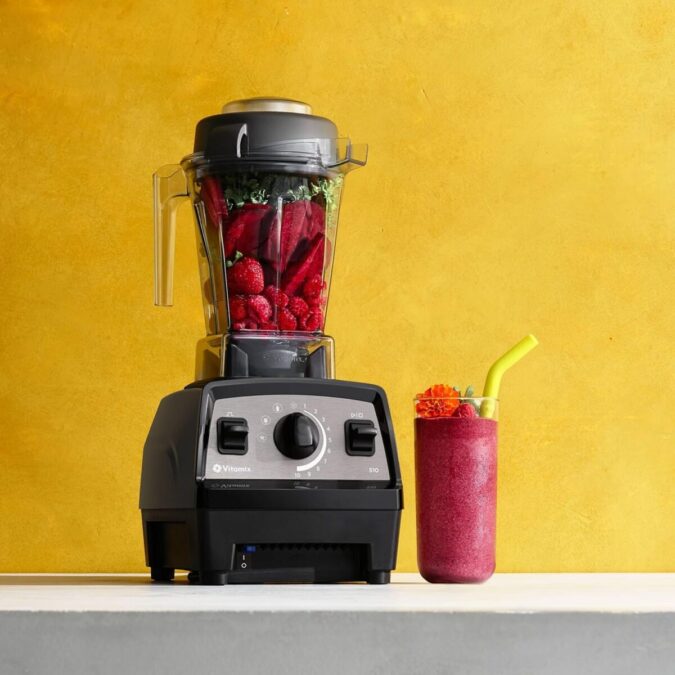 Vitamix Propel 510 blender blending berries and greens with finished smoothie on counter against yellow wall