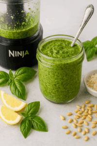A jar of fresh basil pesto with spoon beside a Ninja food processor bowl