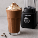 Frozen mocha protein shake in a glass topped with whipped cream beside the Chefman Obliterator blender
