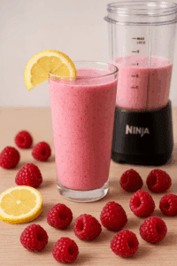 Pink raspberry protein shake in a glass with lemon slices and fresh berries beside a Ninja BN401 cup