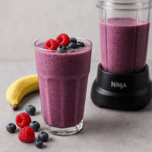 Triple berry smoothie in a tall glass with raspberries, blueberries, and banana beside a Ninja Compact blender cup